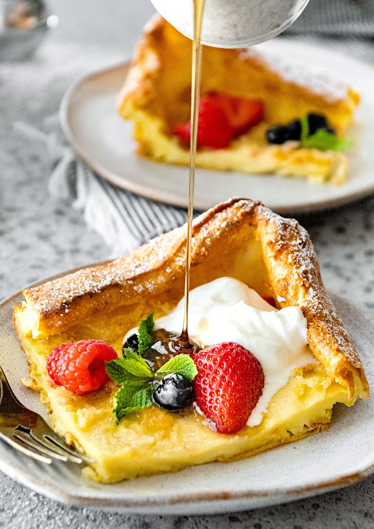 maple syrup being poured over a German pancake with berries and whipped cream. 