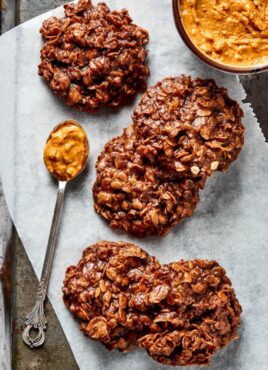 Five no bake cookies on a piece of parchment paper, next to a bowl of peanut butter and a spoonful of peanut butter