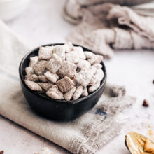 A bowl of puppy chow on a grey cloth, surrounded by scattered puppy chow and a spoon covered with peanut butter.