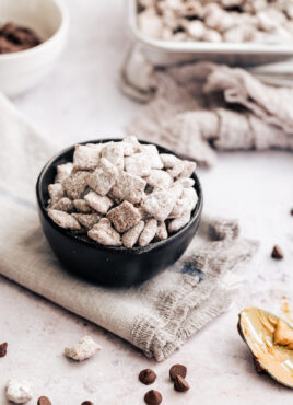 A bowl of puppy chow on a grey cloth, surrounded by scattered puppy chow and a spoon covered with peanut butter.