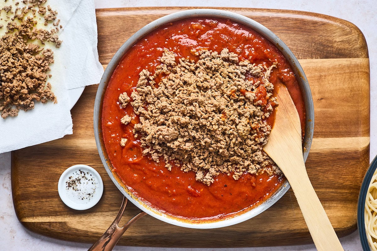 ground beef being stirred into marinara sauce in a skillet. 