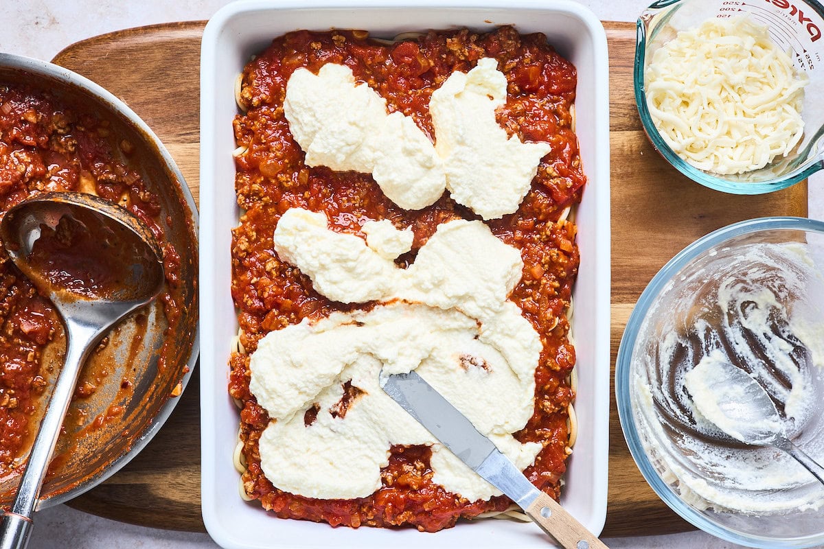 baked spaghetti being layered in baking dish with pasta, meat sauce, and cheese mixture. 