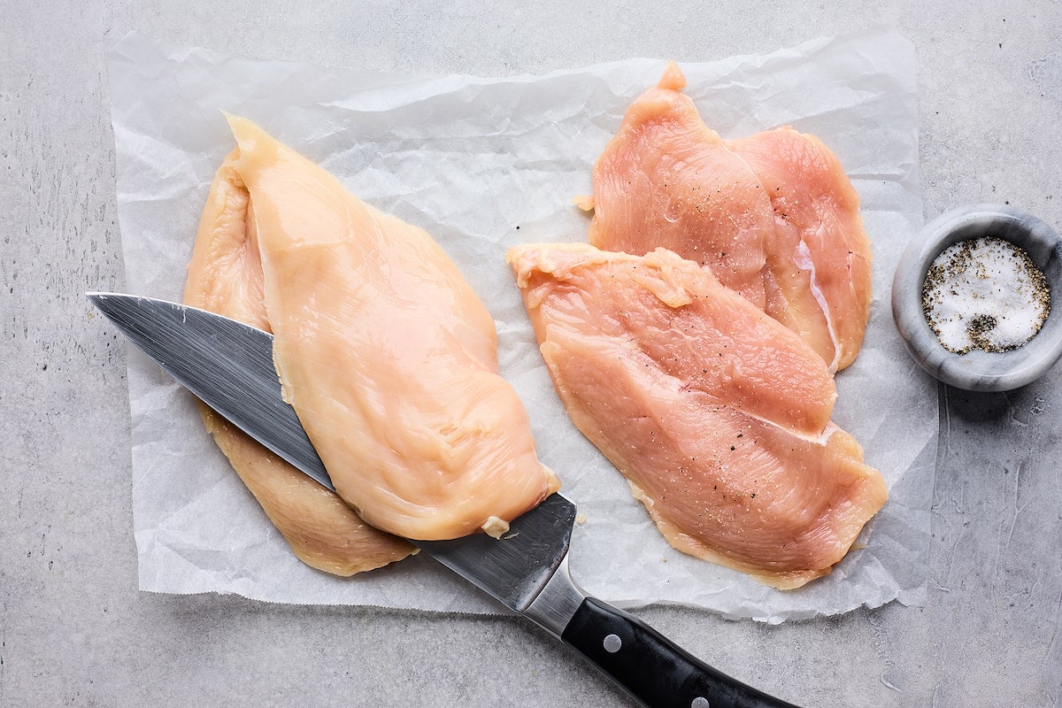 boneless skinless chicken breasts being cut in half with sharp knife to make spinach artichoke chicken. 