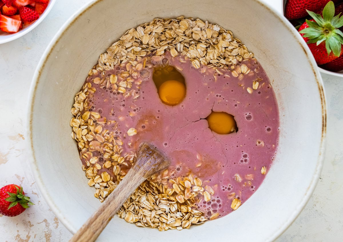 oats, eggs, and strawberry milk in mixing bowl with wooden spoon to make strawberry baked oatmeal. 