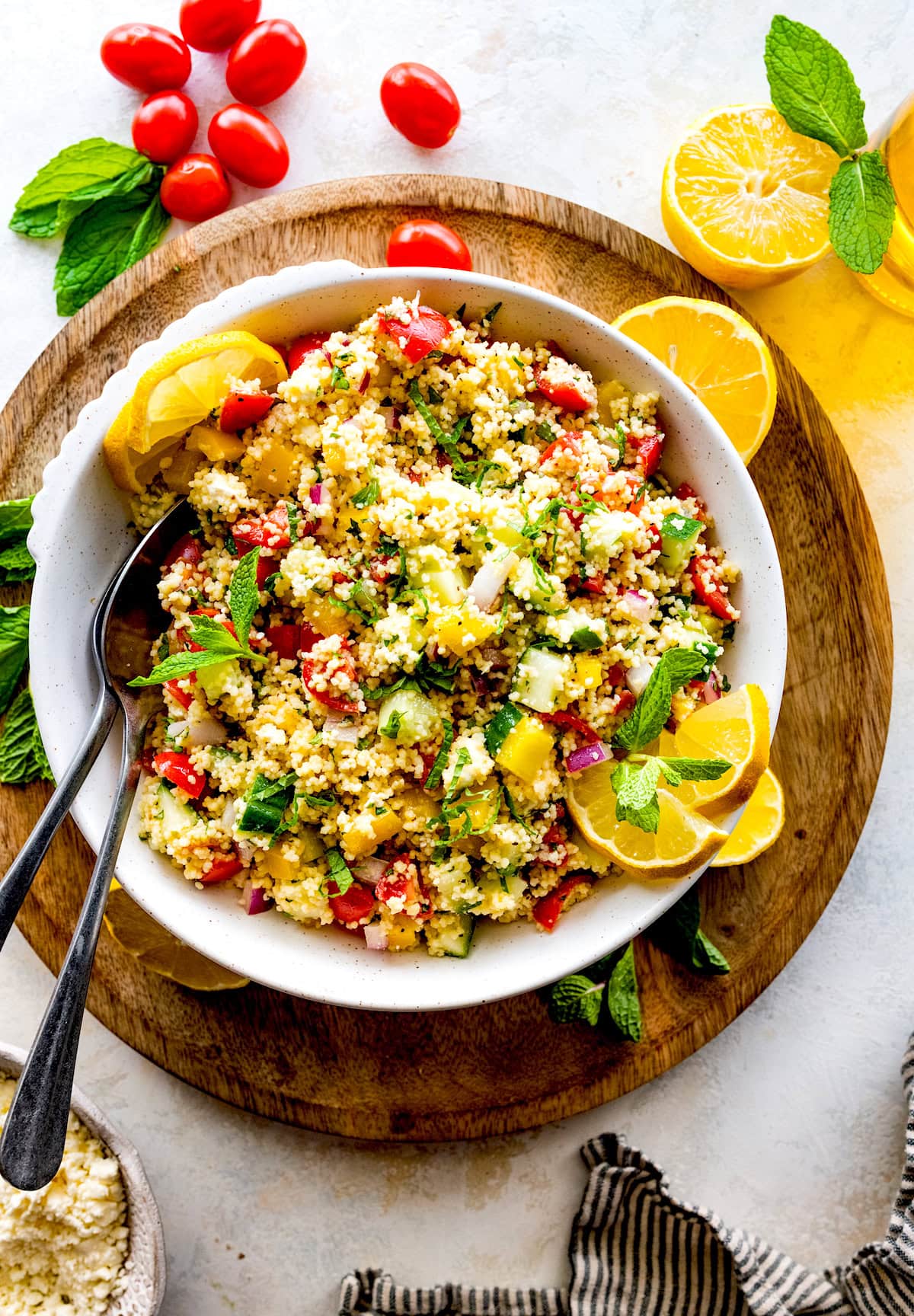 couscous salad with tomatoes, cucumbers, peppers, red onion, herbs, and lemon wedges in white bowl with serving spoons. 