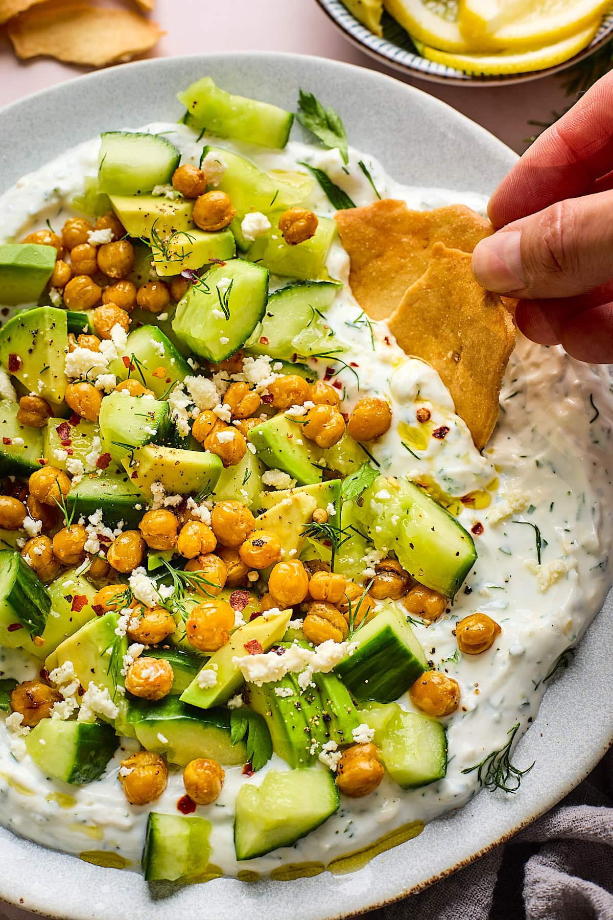 smashed cucumber, avocado, crispy chickpea salad with creamy yogurt feta spread on plate with pita chips being dipped. 