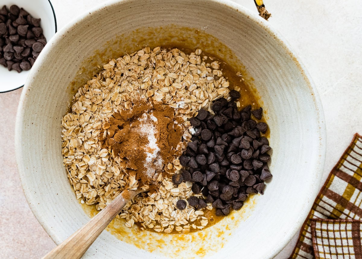 oats, cinnamon, baking powder, salt, and chocolate chips being added to wet ingredients in mixing bowl. 