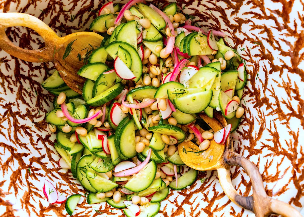 white bean cucumber salad being tossed in bowl with salad servers. 