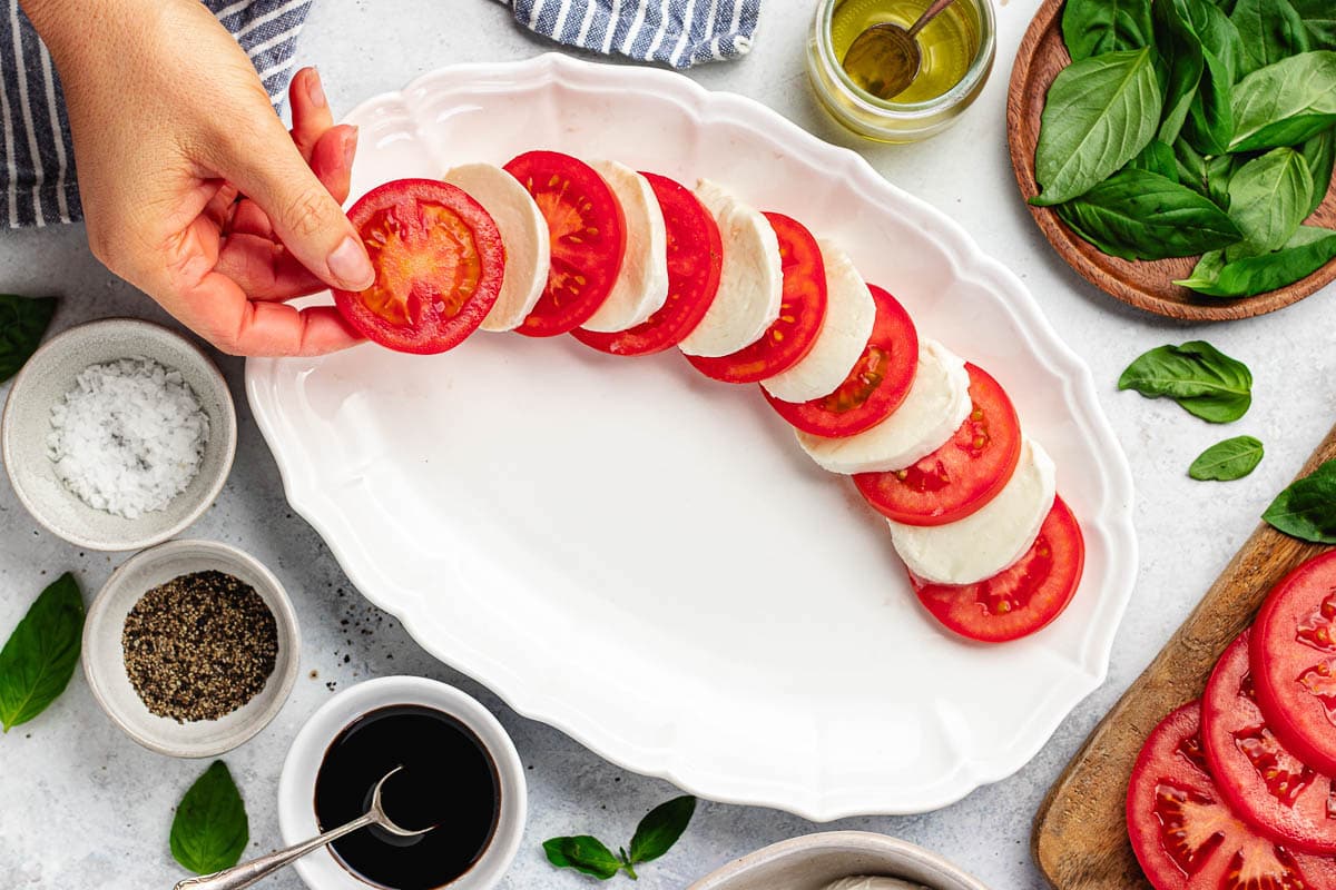 arranging tomato slices and fresh mozzarella slices on large white platter to make caprese salad. 