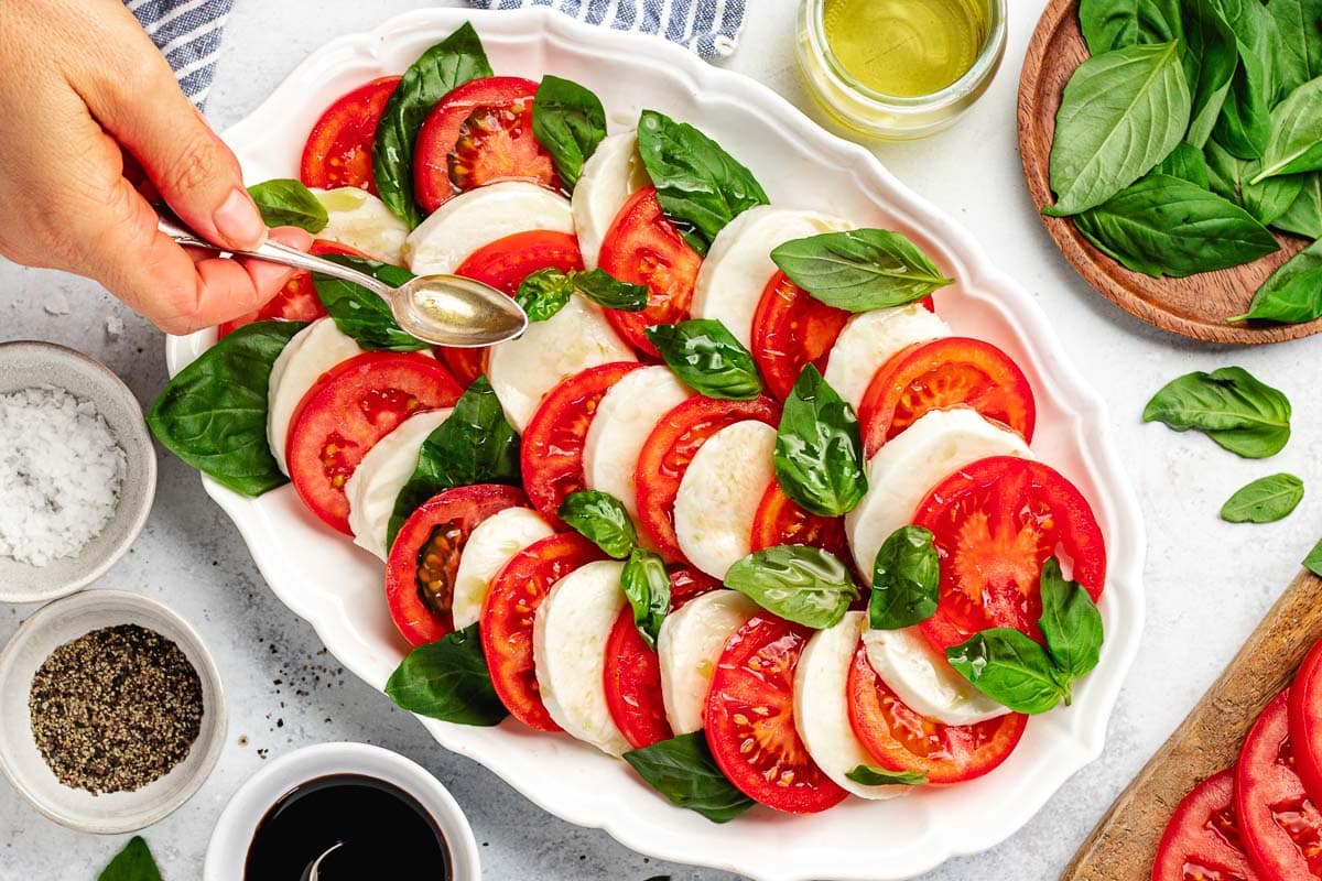 drizzling olive oil over caprese salad on large white platter. 