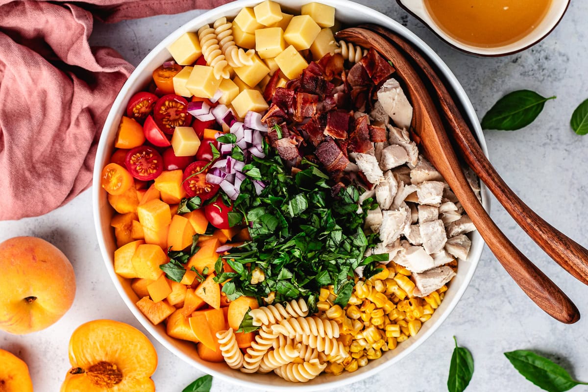 chicken, bacon, white cheddar cheese, tomatoes, peaches, pasta, corn, and basil in big bowl with salad servers. 