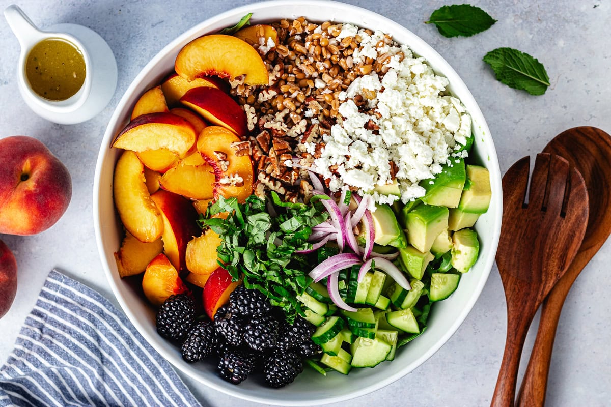 peach slices, pecans, farro, feta cheese, avocado, red onion, blackberries, arugula, herbs, and arugula in bowl. 