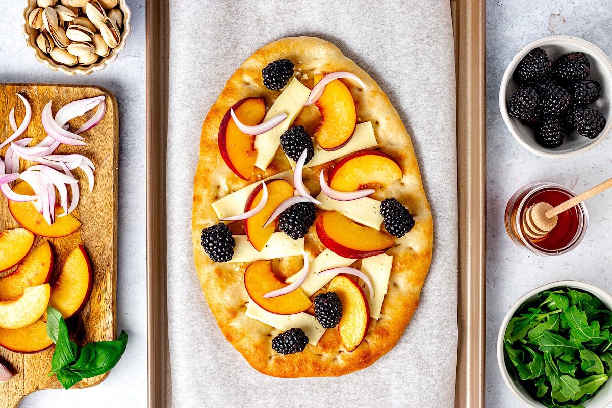 flatbread with brie, peaches, blackberries, and red onion on baking sheet with parchment paper. 