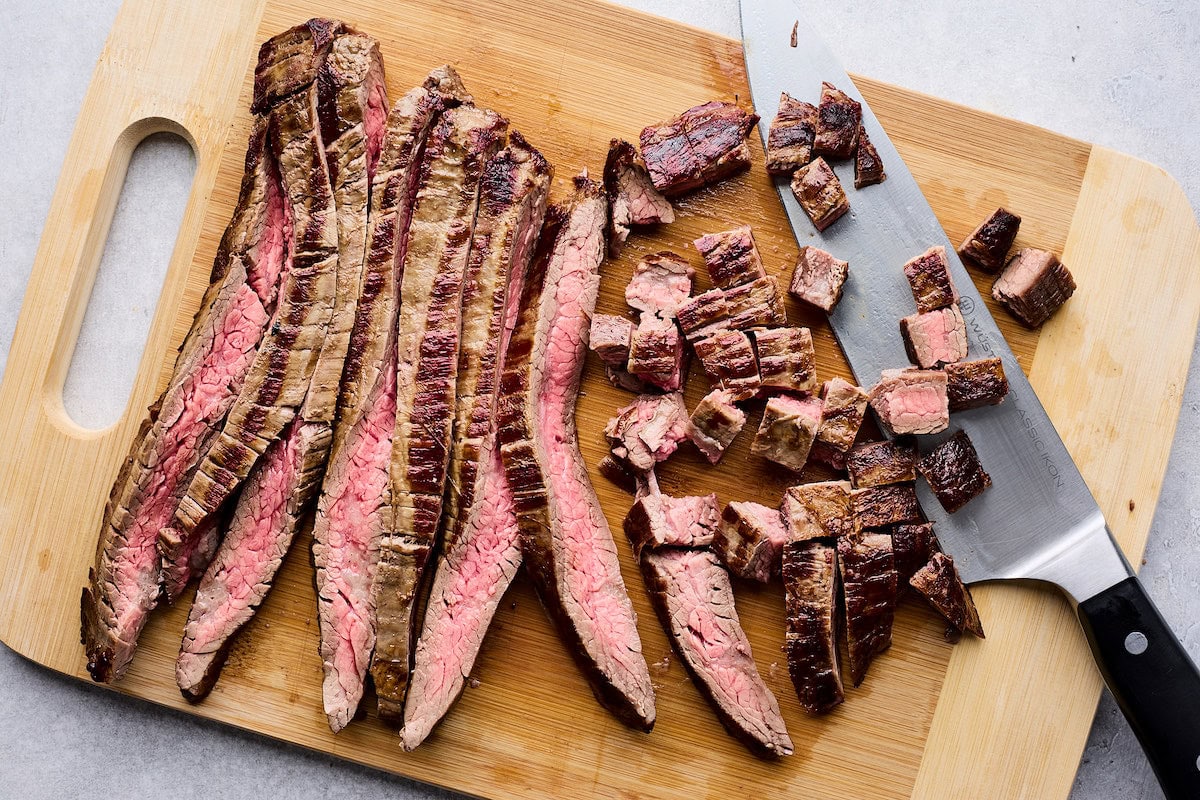 steak being cut into cubes with knife on wood cutting board. 