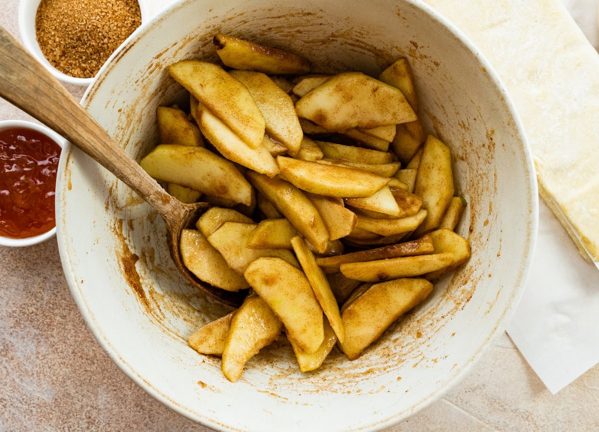 apple slices tossed in sugar and cinnamon in mixing bowl with wooden spoon. 