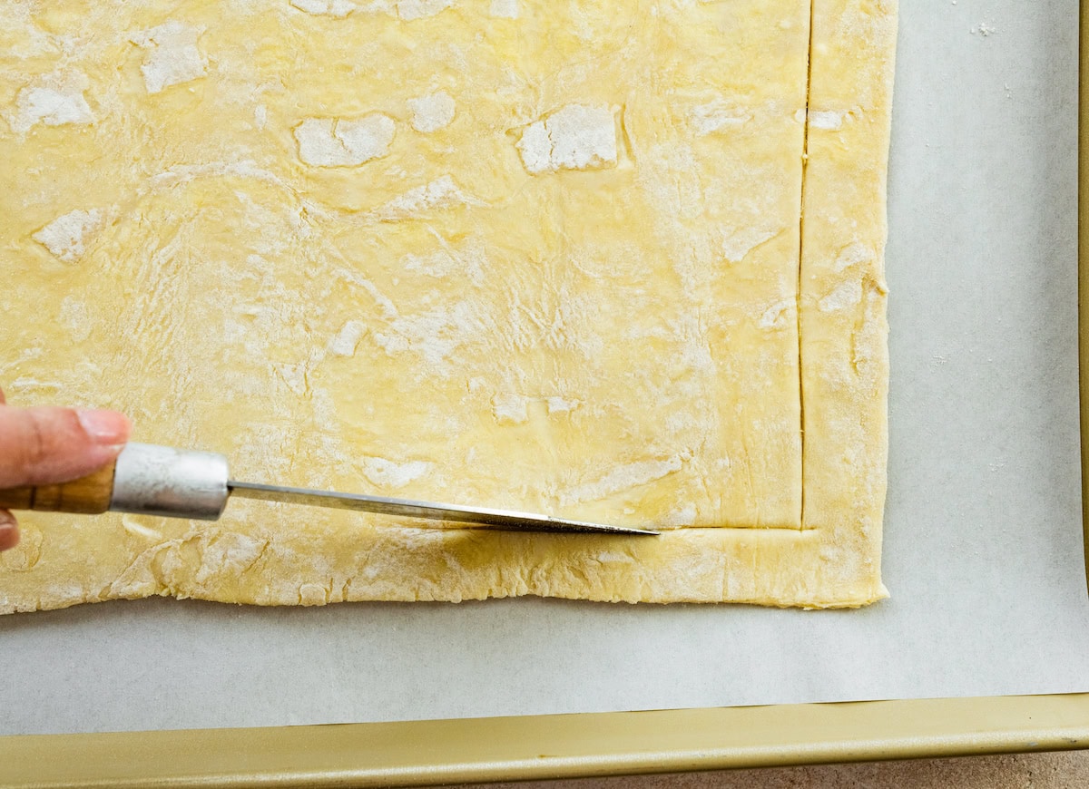 a knife scoring the edge of a puff pastry sheet to make an apple tart. 