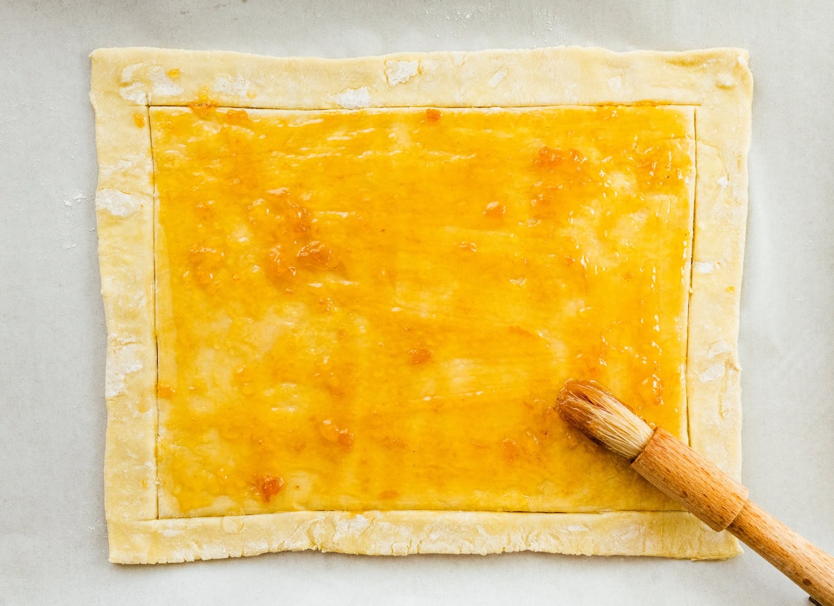 apricot preserves being brushed on puff pastry to make apple tart. 