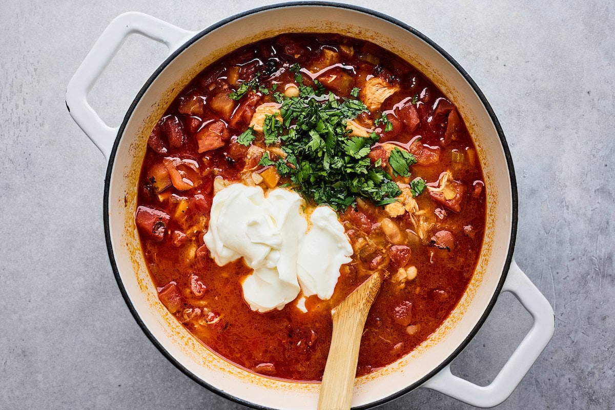 plain Greek yogurt and cilantro being added to buffalo chicken chili in white pot. 