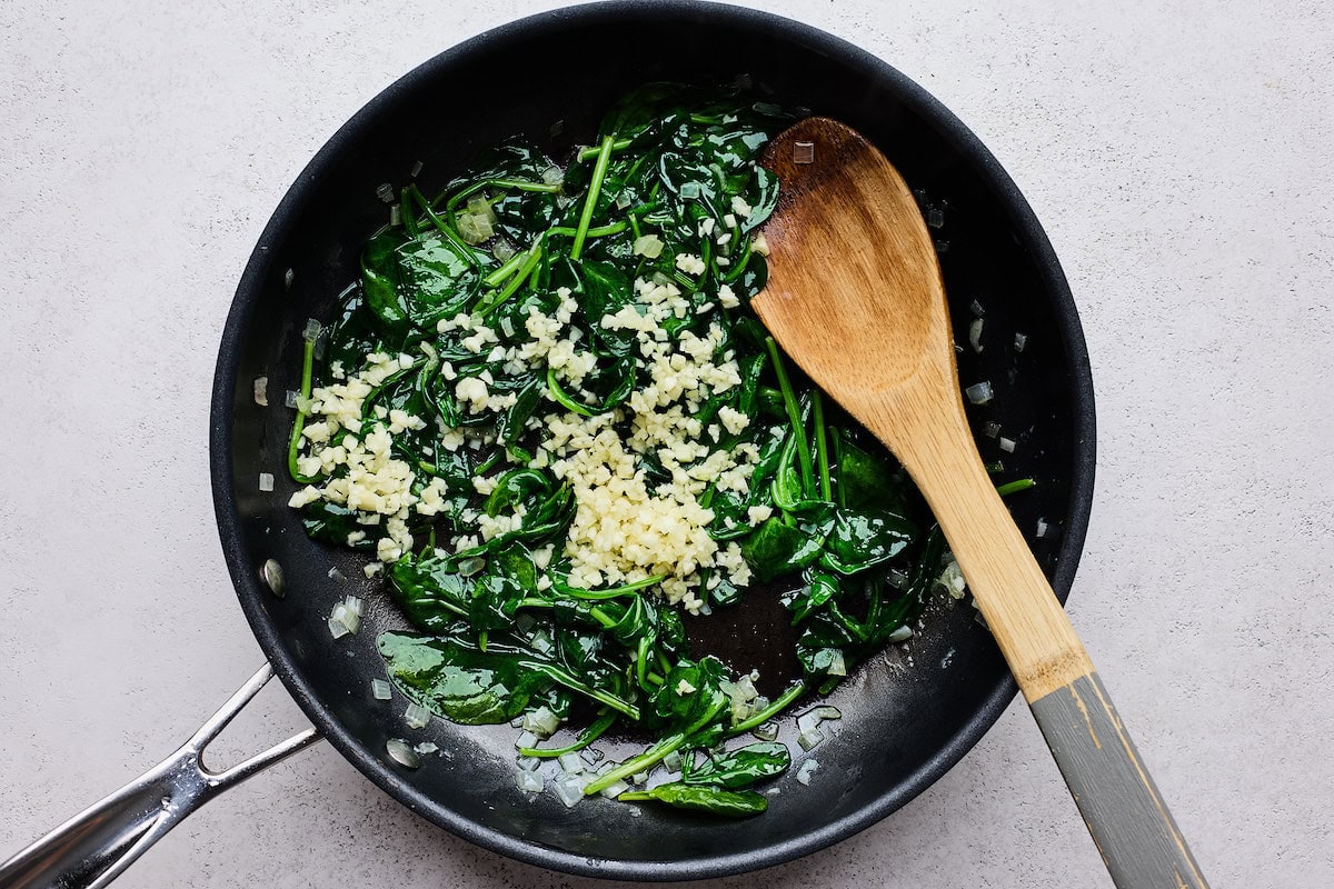 shallot, spinach, and garlic cooking in skillet with wooden spoon. 