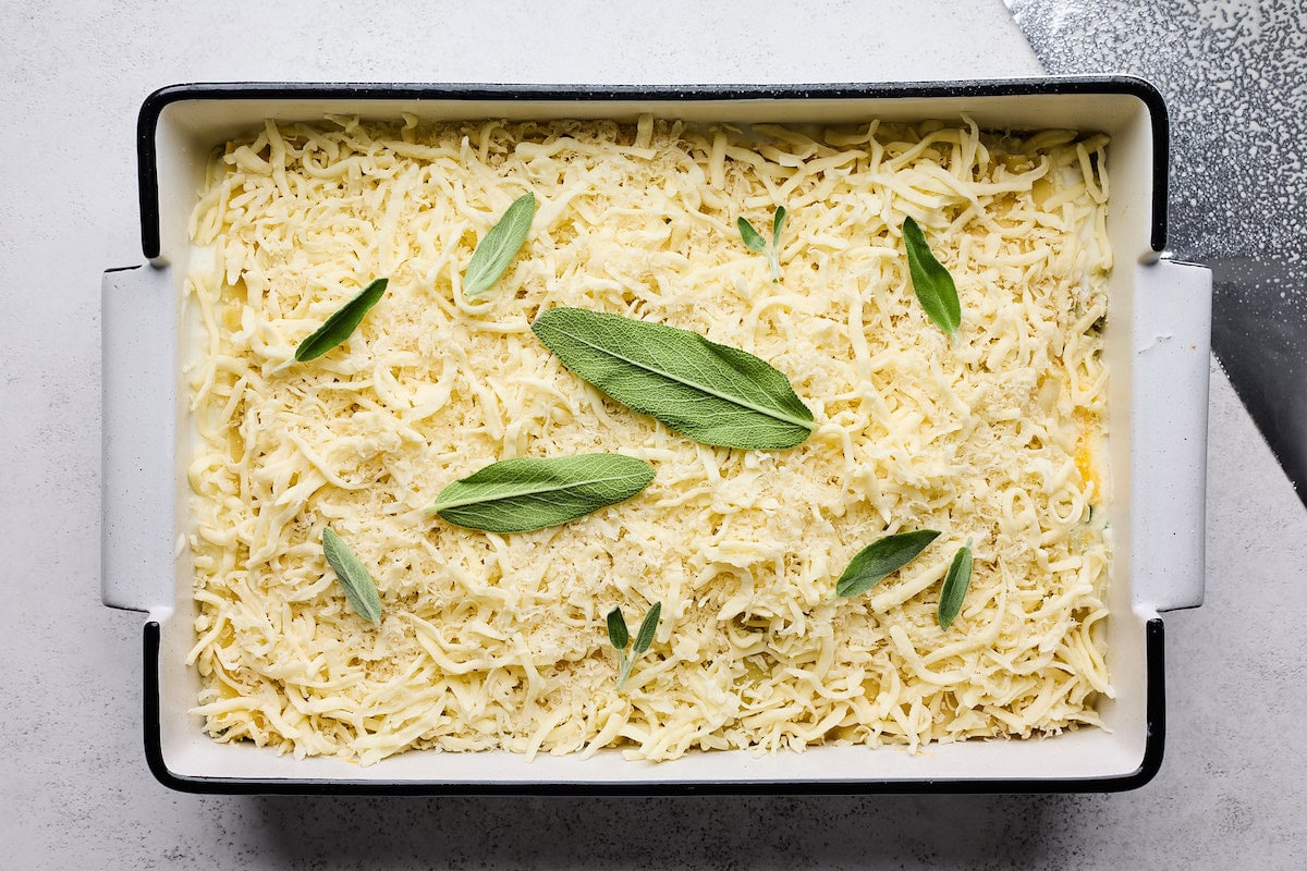 butternut squash being assembled in baking dish with cheese and sage leaves on top. 