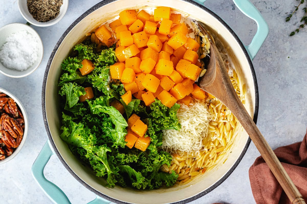 butternut squash, kale, and parmesan cheese being stirred into orzo in a pot. 