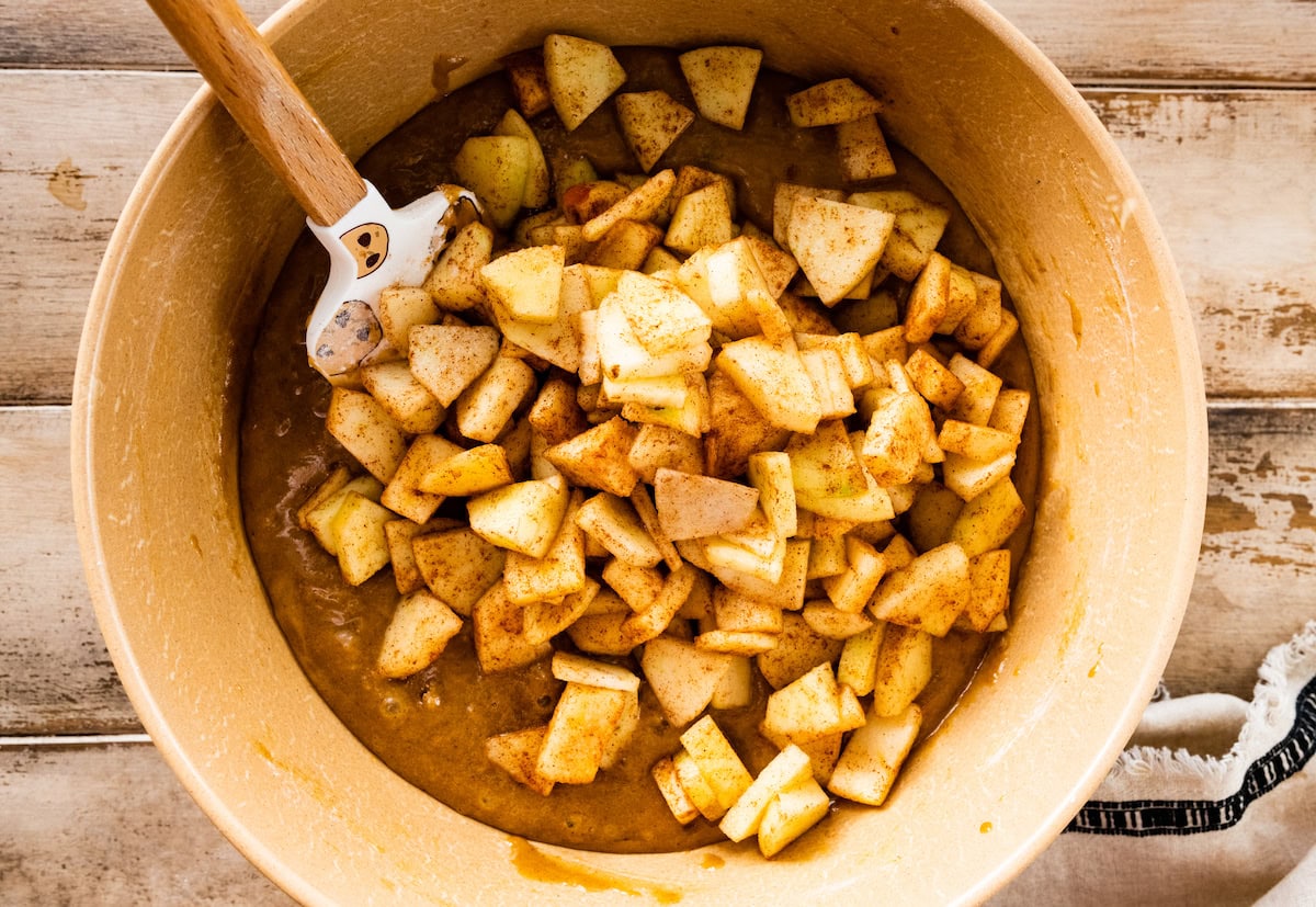 chopped apples being stirred into apple cake batter in mixing bowl. 