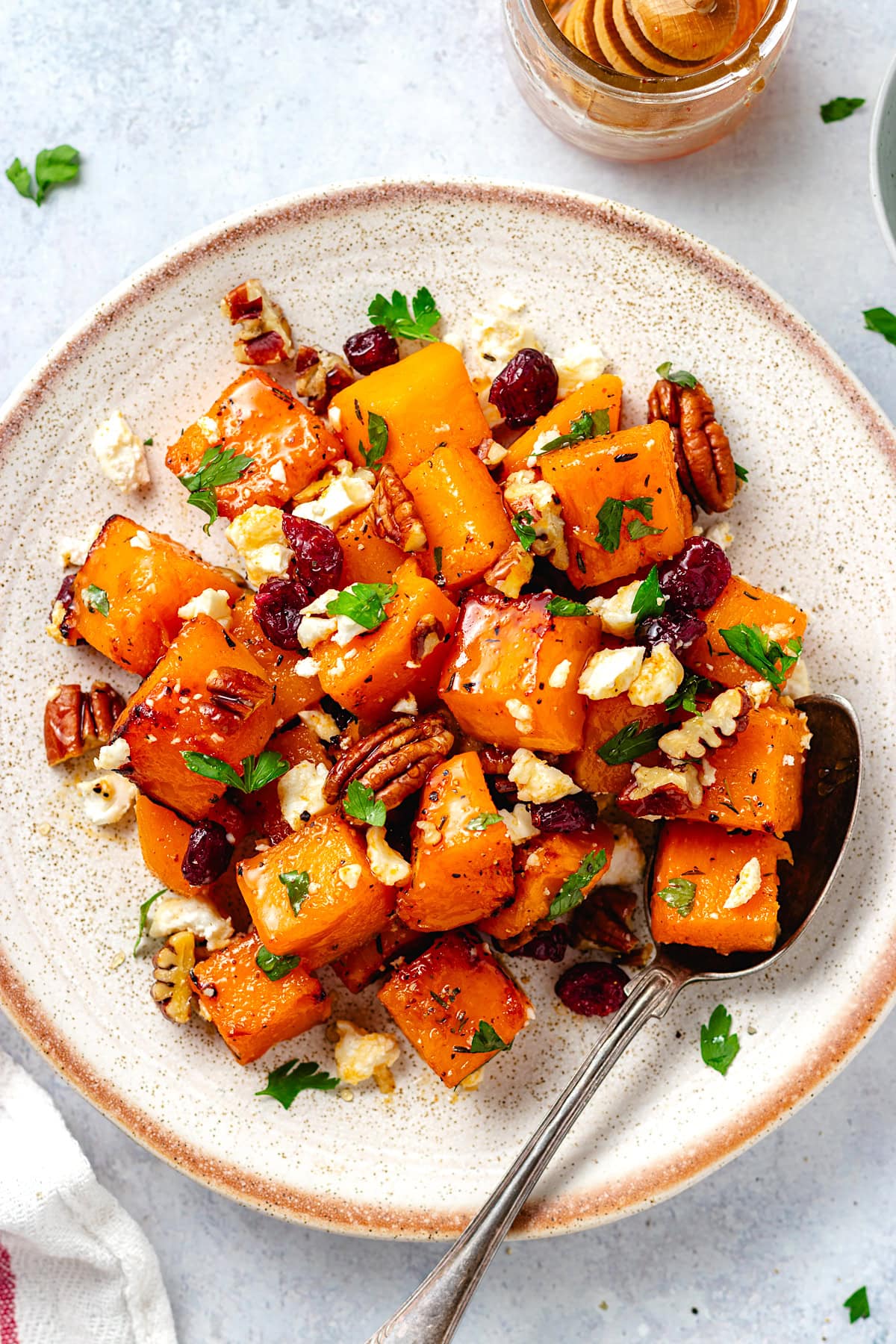 roasted butternut squash with pecans, dried cranberries, feta, parsley, and hot honey on plate with spoon. 