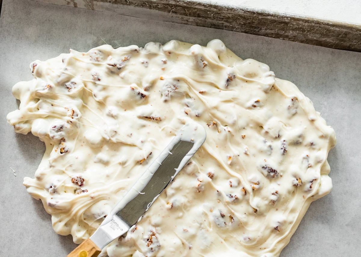 melted white chocolate and candied pecans being spread on baking sheet with parchment paper to make bark. 