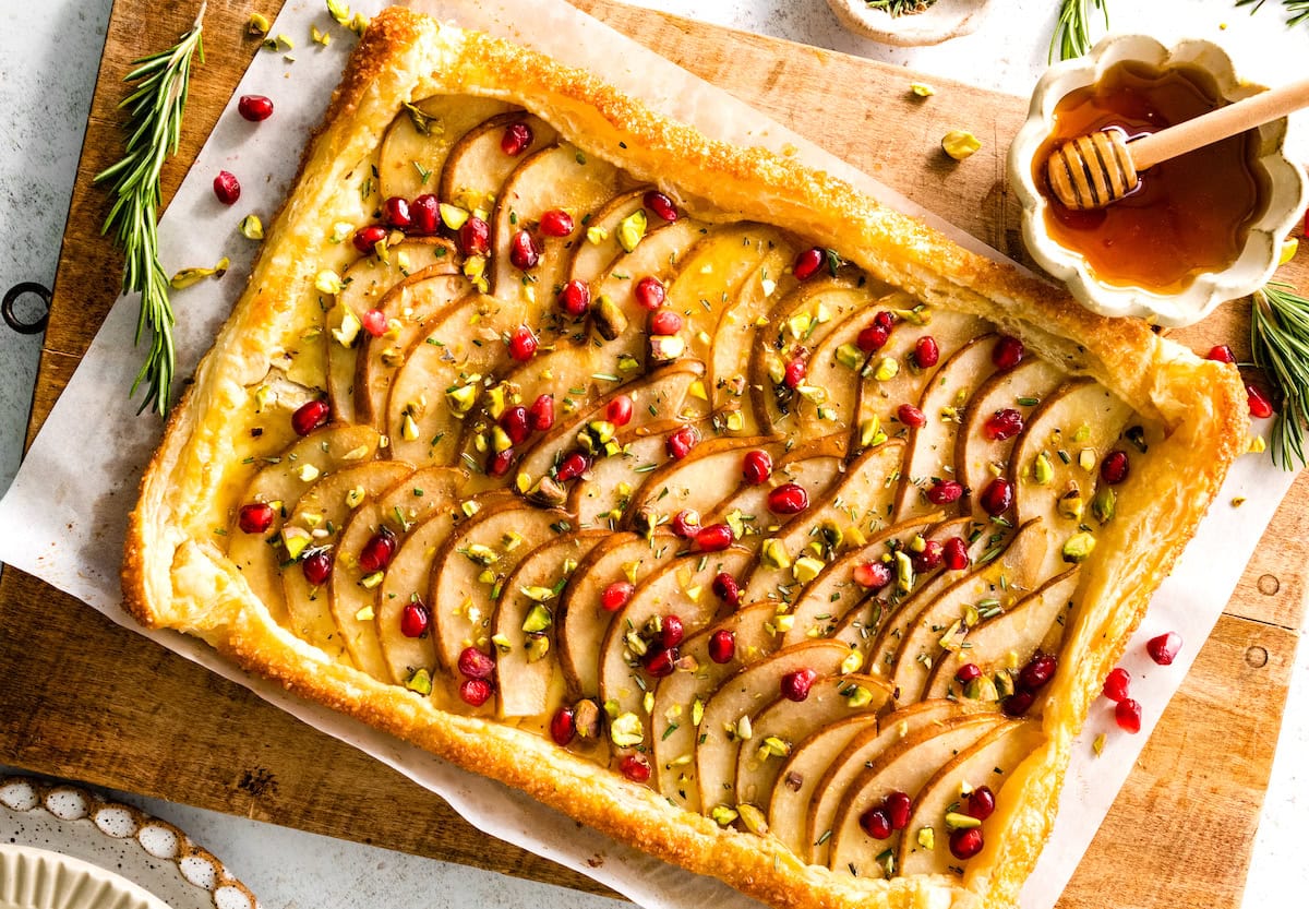 pear brie puff pastry tart with pomegranate, pistachios, rosemary, and honey on wood cutting board. 