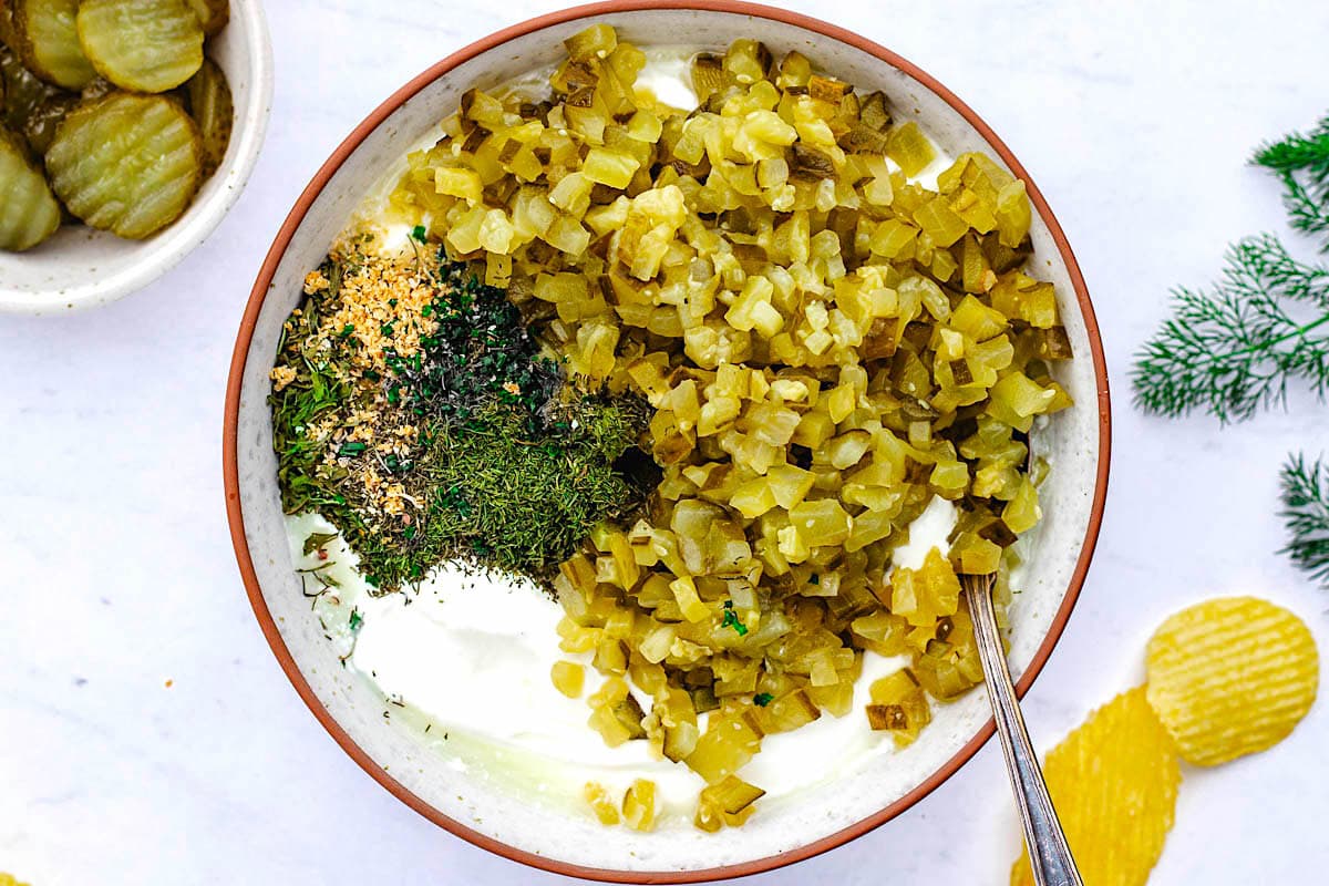 chopped dill pickles, seasonings, herbs, and Greek yogurt in bowl with spoon to make dill pickle dip. 