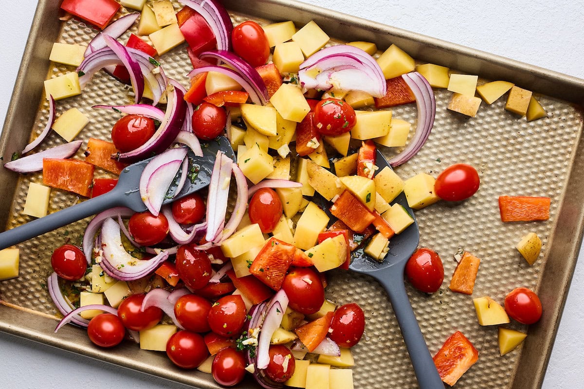 potatoes, grape tomatoes, red bell pepper, and red onion being tossed in Greek marinade on sheet pan. 