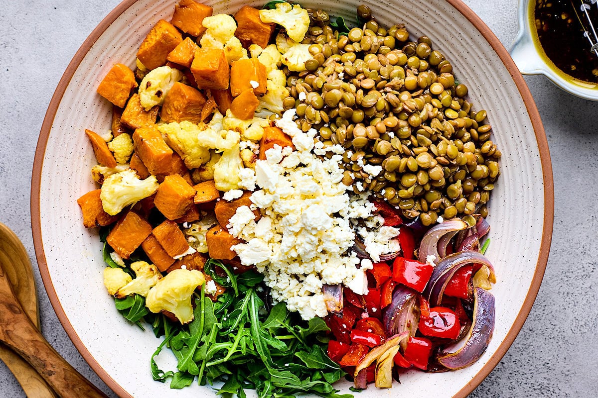 lentils, roasted sweet potatoes, cauliflower, red peppers, red onion, arugula, and feta cheese in bowl. 