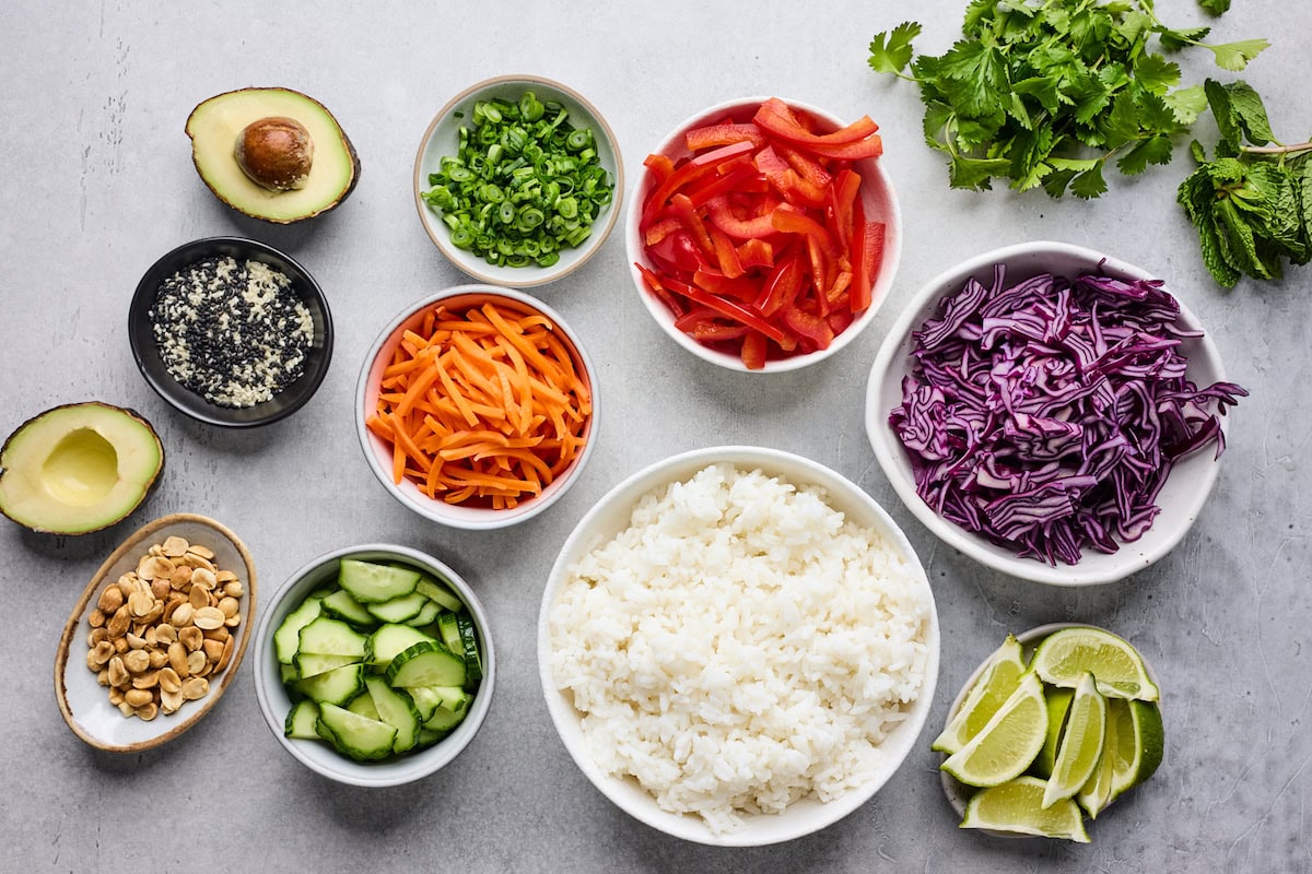 avocado, green onion, red pepper, herbs, carrots, cabbage, sesame seeds, peanuts, cucumber, rice, and lime wedges in bowls to make shredded tofu peanut bowls. 