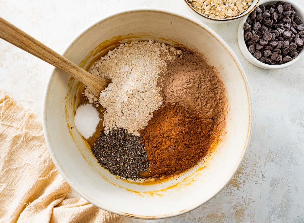 oat flour, cocoa powder, chocolate protein powder, chia seeds, baking soda, and salt being added to mixing bowl with wooden spoon to make healthy chocolate cookies. 