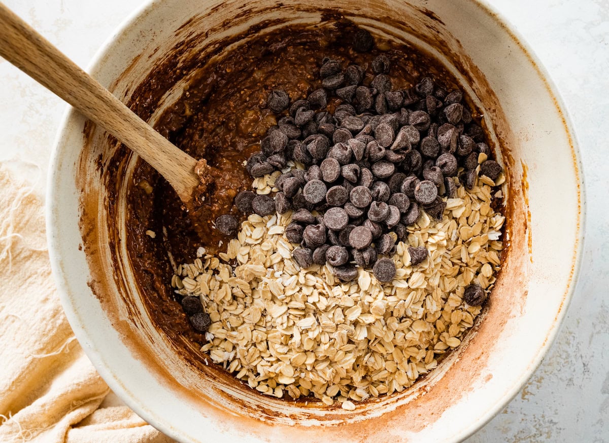 oats and chocolate chips being added to healthy chocolate chip cookie dough in bowl with wooden spoon. 