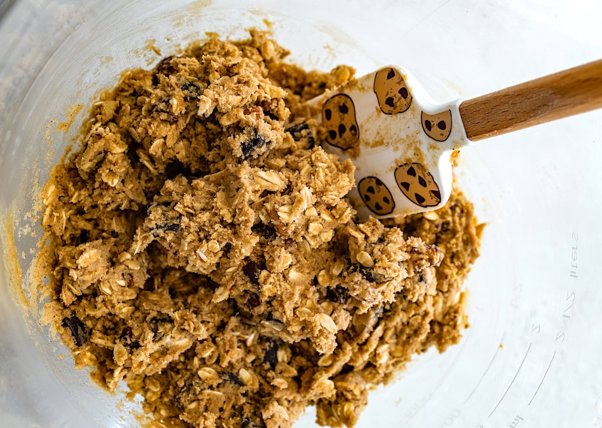 brown butter oatmeal cookie dough with pecans, dates, and chocolate chunks in mixing bowl with spatula. 