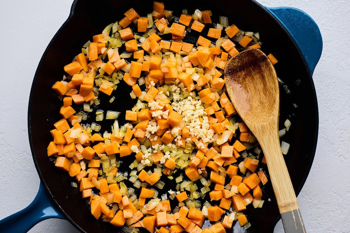 sweet potatoes, onion, and garlic cooking in cast iron skillet with wooden spoon. 