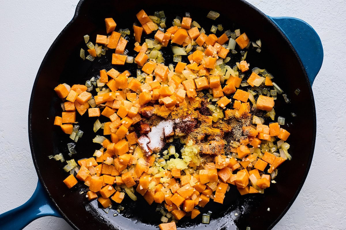 spices being added to sweet potatoes, onion, and garlic cooking in cast iron skillet. 