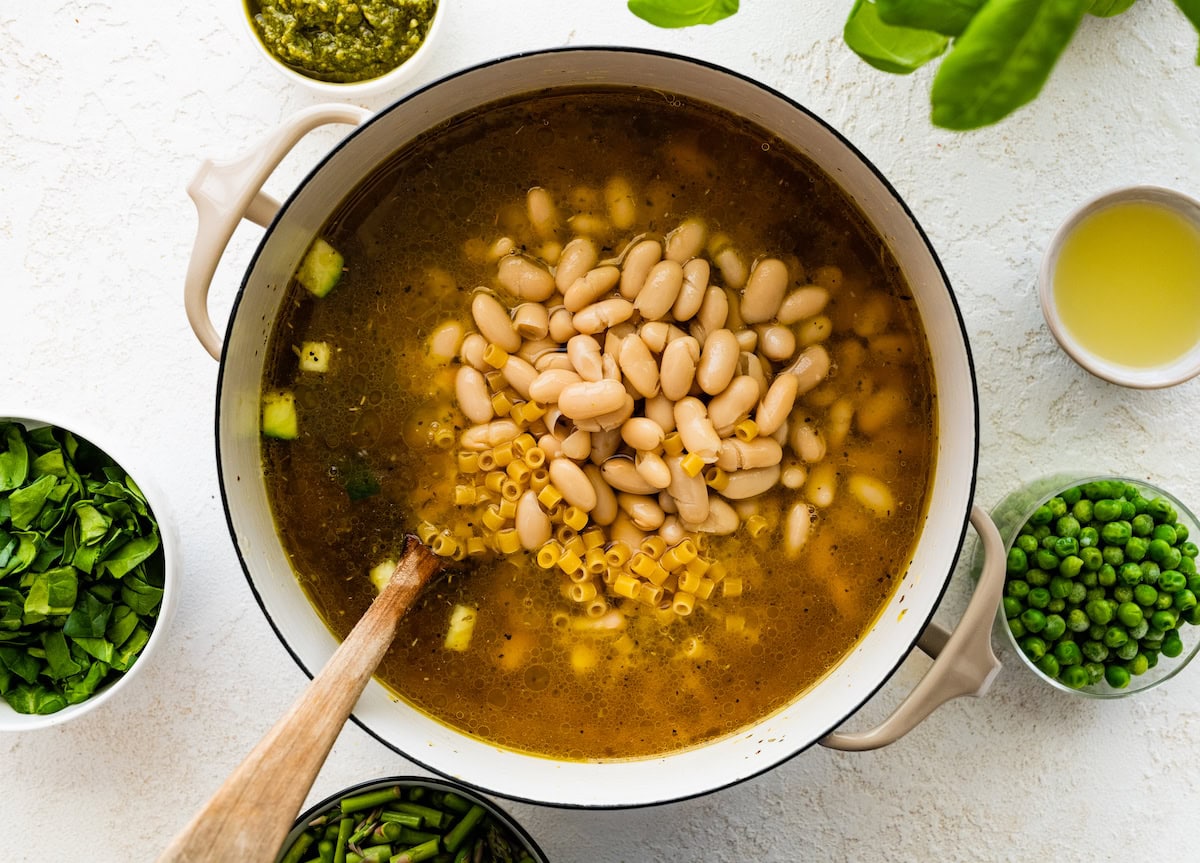 vegetable broth, white beans, and pasta being added to pot to make spring minestrone soup. 